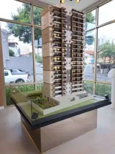 Maquete de um edifício residencial alto, exibida em uma base de vidro em um ambiente interno.