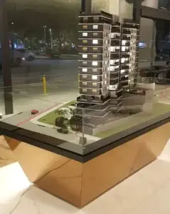 Maquete iluminada de empreendimento imobiliário com duas torres e área de paisagismo, em uma base moderna de vidro e metal.